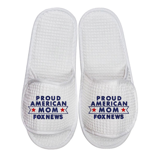 A pair of white waffle-knit slippers featuring the FOX News Proud American Mom logo embroidered in red, white, and blue, displayed in a bright American home setting.