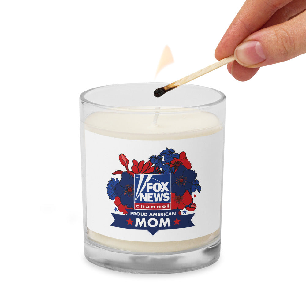 A FOX News Proud American Mom candle displayed on a mantle next to a small American flag in a patriotic home setting.
