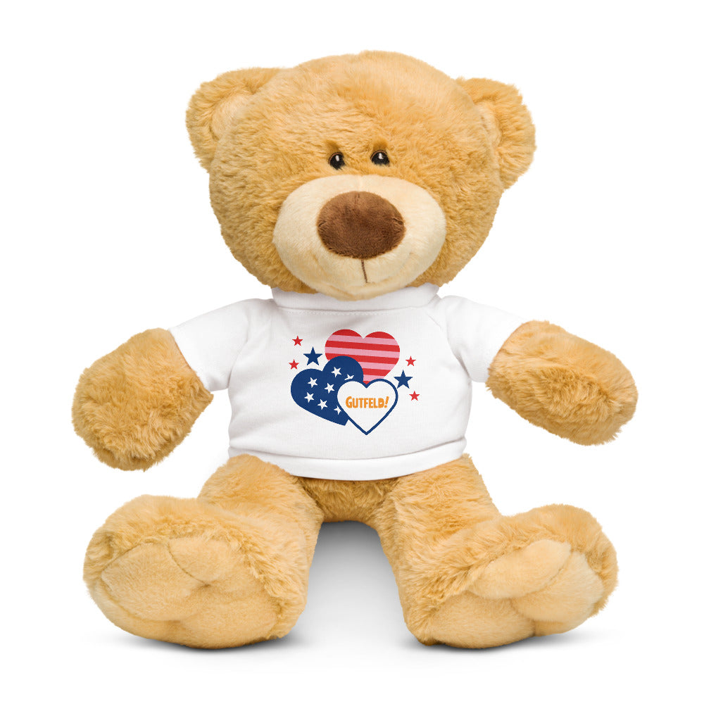 A soft brown teddy bear with a white heart graphic on its chest featuring the Gutfeld! logo, sitting in a comfortable American home office.