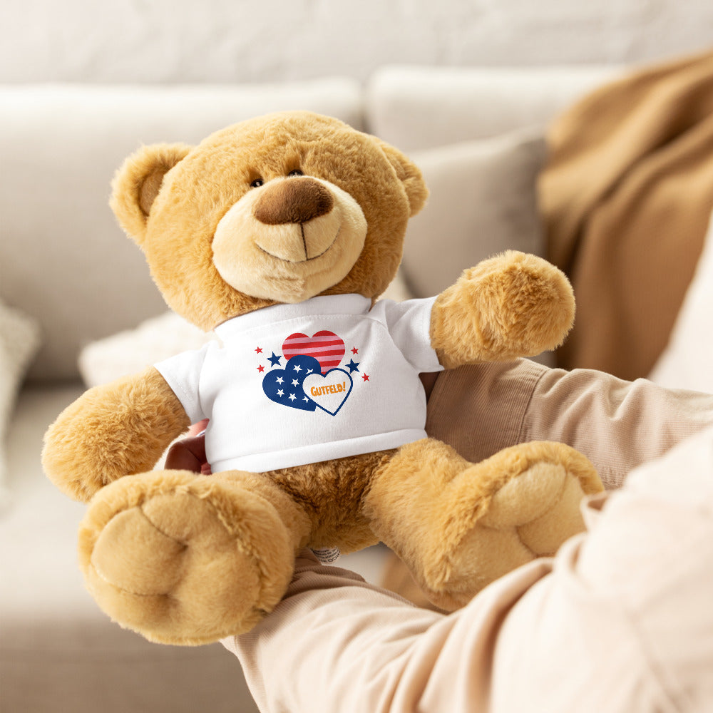 The Gutfeld! Hearts Teddy Bear displayed on a clean shelf next to other Fox News collectibles.