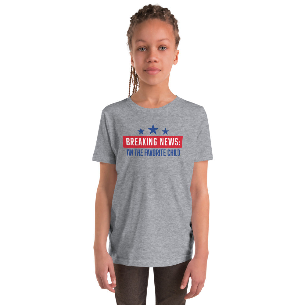 A young boy sitting in a clean American living room wearing the FOX News Breaking News Kids T-Shirt.