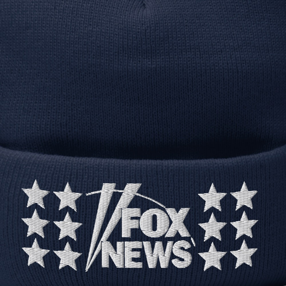 A red FOX News pom pom beanie featuring a crisp white embroidered logo, shown on a wooden table near a window.